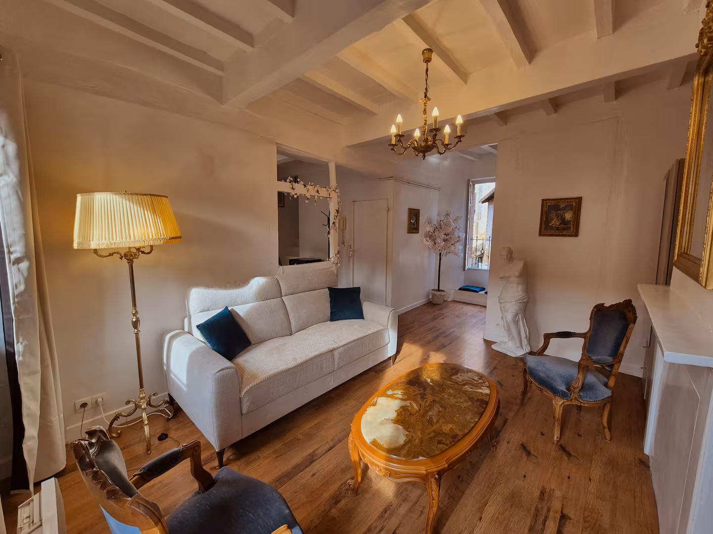Le salon d’un logement à Albi : canapé clair, lustre, plafond à poutres blanches